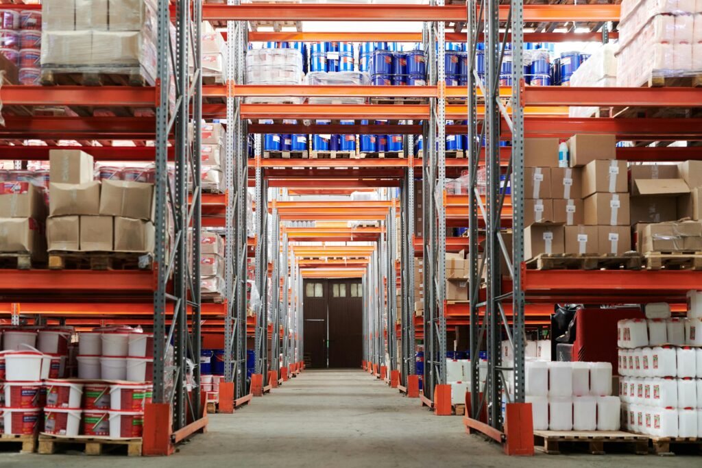 Wide interior view of a large warehouse aisle with tall orange metal racks stacked with boxes, pallets, and containers on both sides.